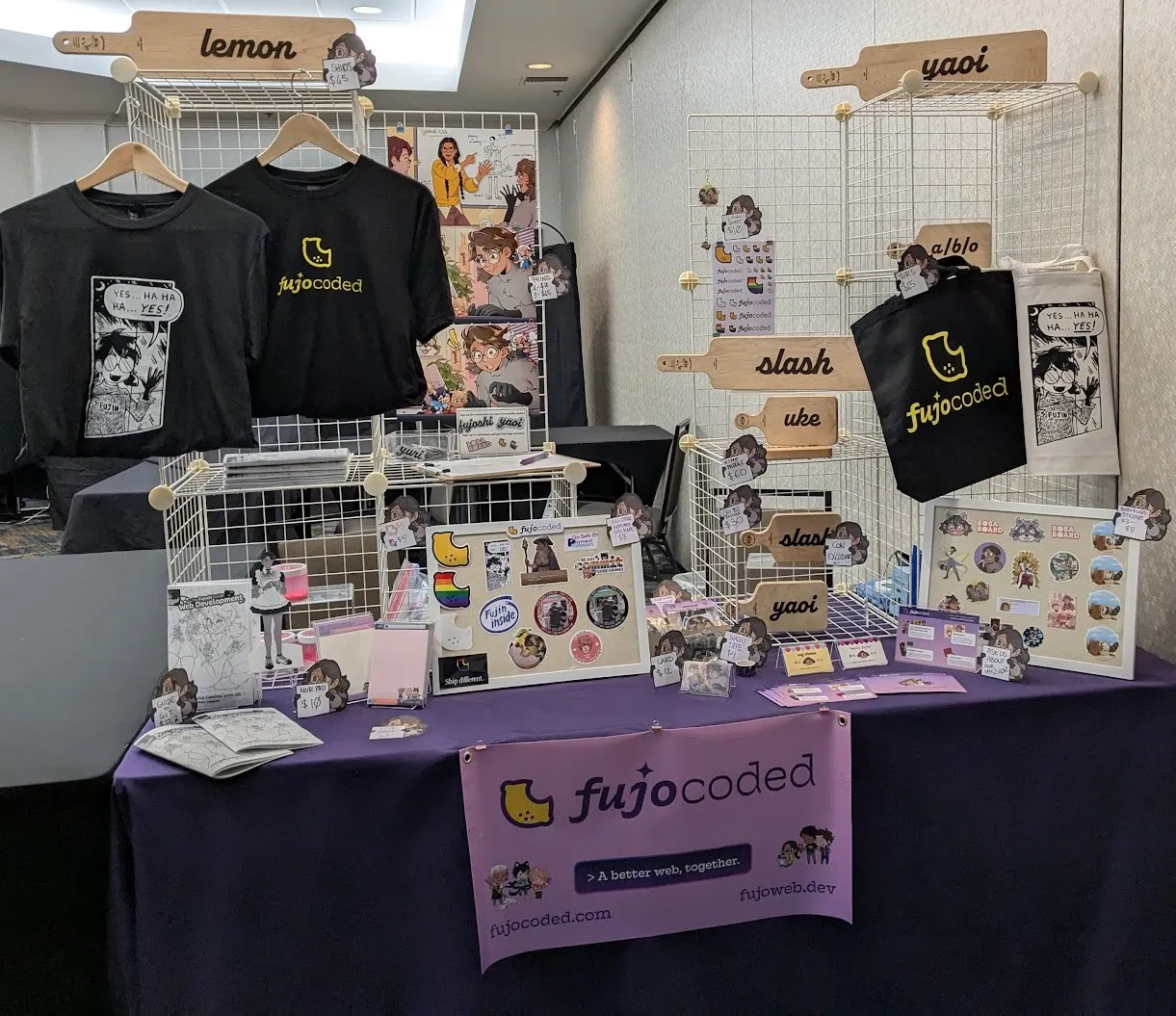 Photo of a dealer's table in a hotel meeting room. A small professionally printed banner with the FujoCoded logo and the slogan "A better web, together" hangs on the front of the table, which is covered in a purple tablecloth. Cream-colored wire cubes in 3 by 2 stacks are arranged on either end of the table to create surfaces for merchandise. On the left cube stack are Fujin Sickos t shirt, a FujoCoded lemon logo shirt, 3 FujoCoded art prints, a newsletter signup sheet offering free stickers in exchange for signup, several containers of catboy bathwater, and a hime-size cheese paddle that says 'lemon'. On the right cube stack are several hime and chibi size cheese paddles, a Fujocoded logo tote bag and a Fujin sickos tote bag, some phone charms, and a page of FujoCoded logo stickers. In front of the left cube is an array of printed-out FujoGuides with a standee of HTML as a paper doll wearing a maid outfit. In the middle front of the table is a small bulletin board offering an array of FujoCoded stickers from the FujoCoded kickstarter. On the right side of the table is an array of business cards and info cards about FujoCoded's different projects, followed by another small bulletin board of older BobaBoard stickers.
