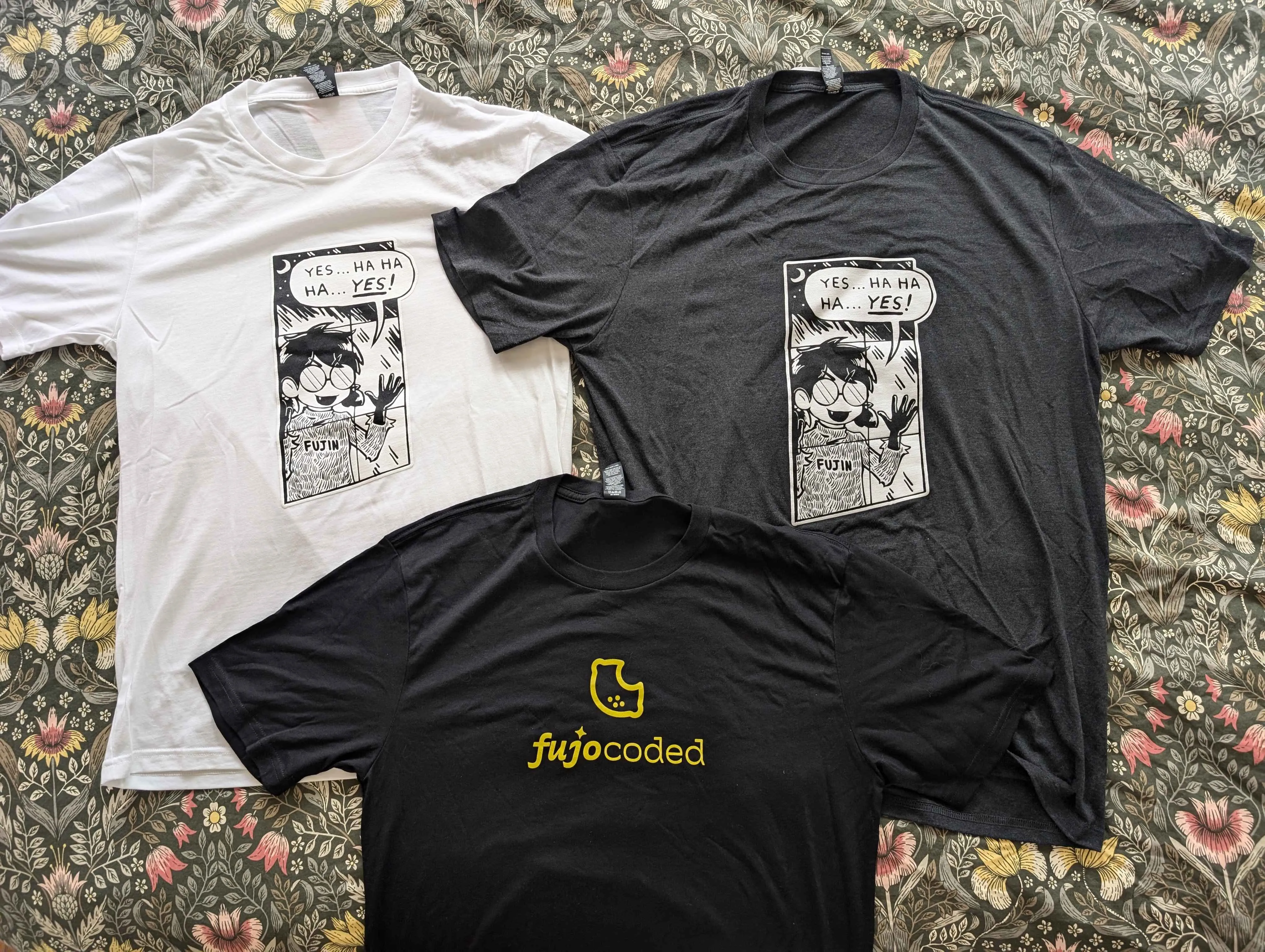 Three tee shirts laid flat. The white and grey shirts have the “Yes…Ha Ha
Ha…Yes!” design with Boba-tan pressing her face to a window, while the black
shirt has a bright yellow FujoCoded logo.