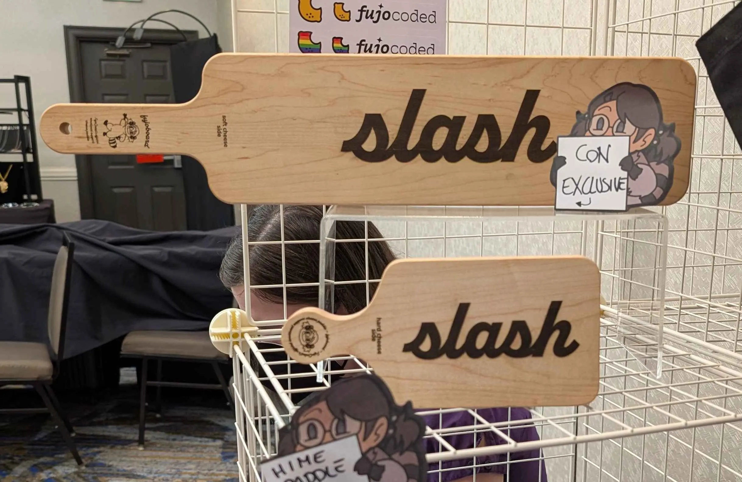 Photo of a large hime-size cheese paddle emblazoned with the word "slash" in lowercase cursive on a stand behind a smaller chibi-size cheese paddle with the same design. A post-it note of Boba-tan holding a white square has the words "Con Exclusive" with an arrow pointing toward the paddles. A person in a purple shirt and black KN95 mask is obscured behind the paddle display. (That's Dove, ignore her.)